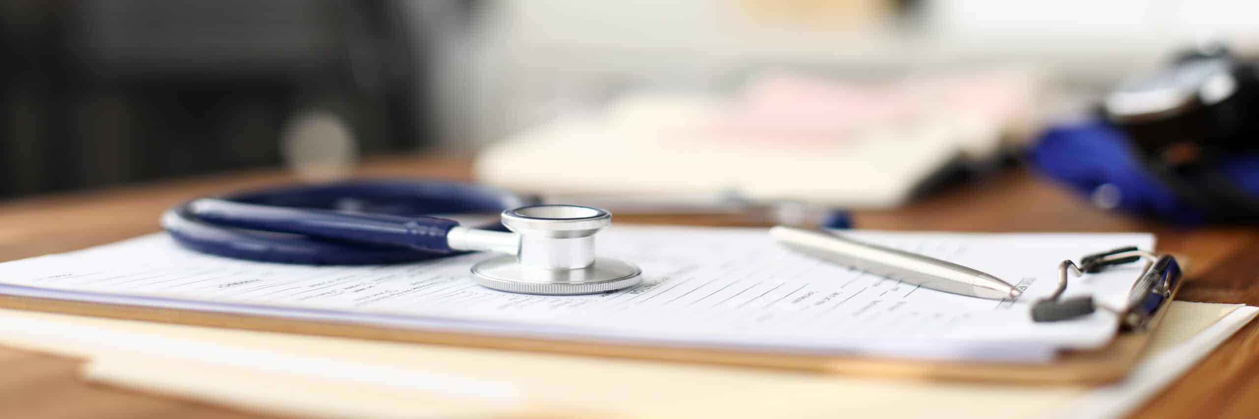 A stethoscope and pen rest on a medical form attached to a clipboard, with blurred documents and a notebook in the background on a wooden desk.