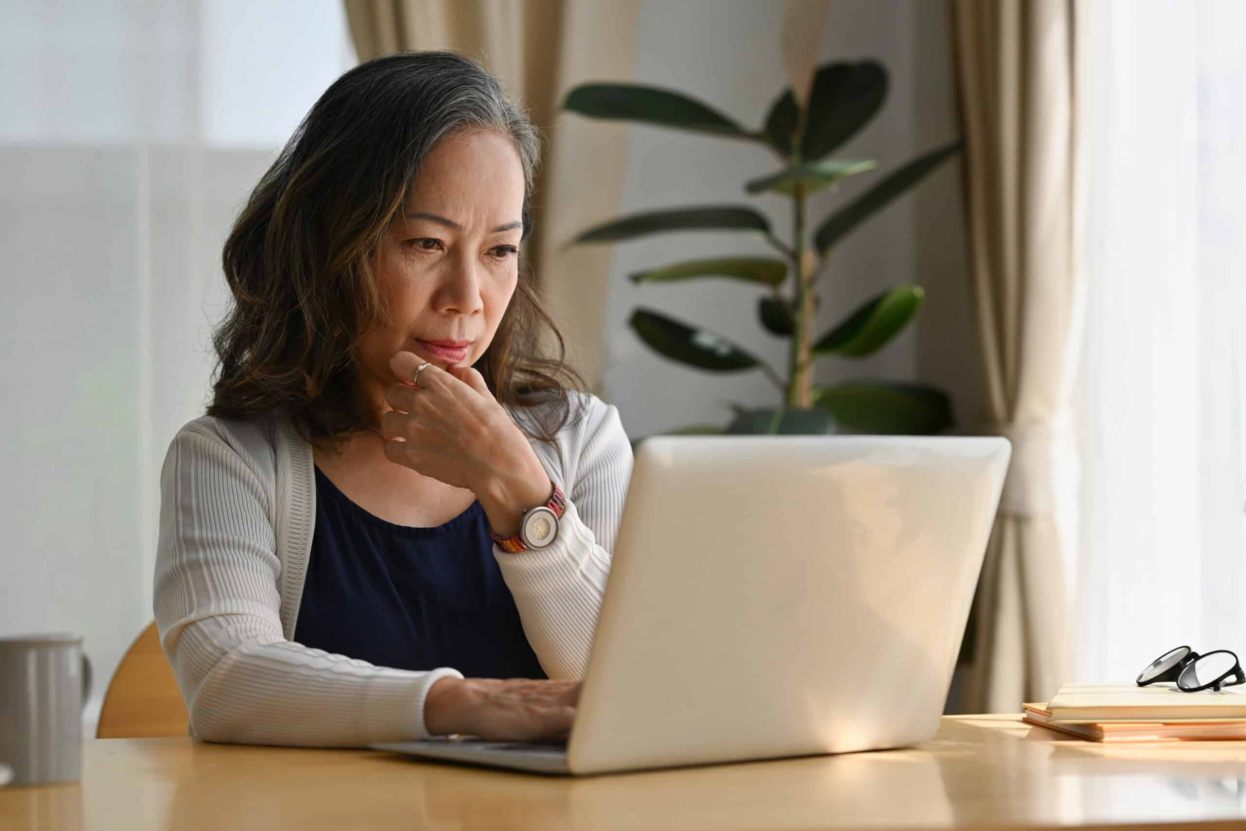 An older woman with long gray hair sits at a table, looking thoughtfully at a laptop screen. She rests her chin on her hand. Papers, a notebook, and glasses are on the table; a potted plant is in the background.