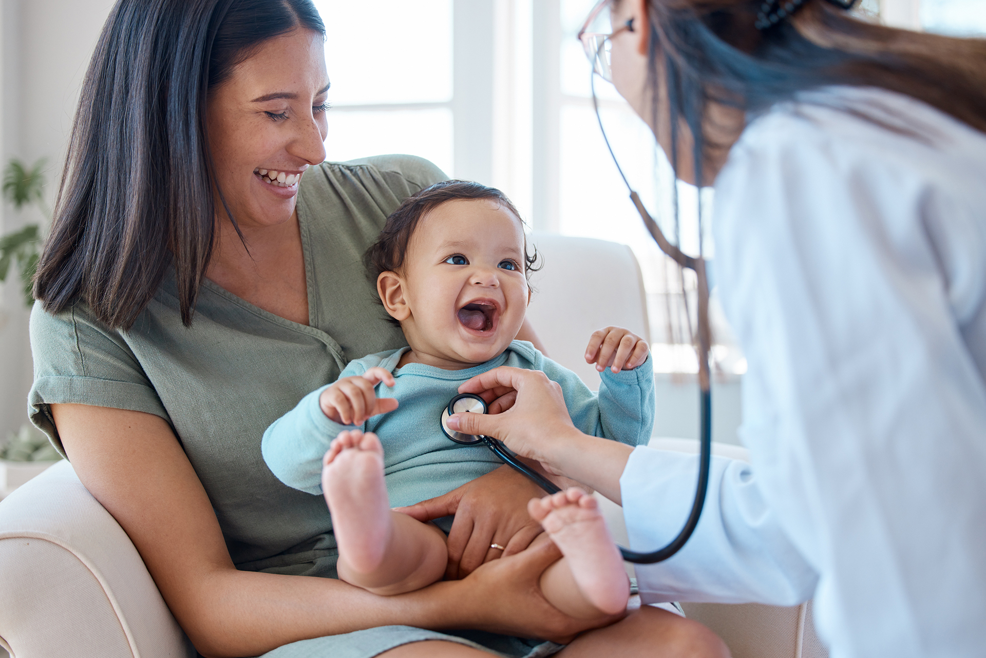 A smiling woman holds a happy baby as a doctor listens to the baby's chest with a stethoscope during a cheerful medical checkup.