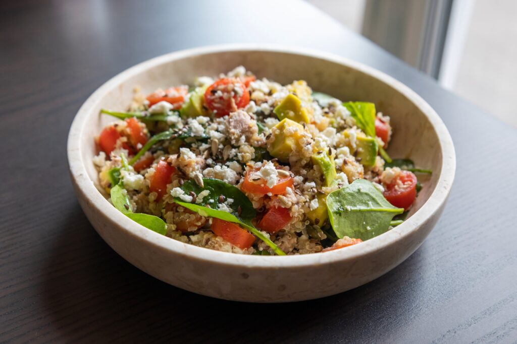 A ceramic bowl of quinoa salad with spinach, tomatoes, avocado, crumbled cheese, and grains on a dark wooden table by a window.