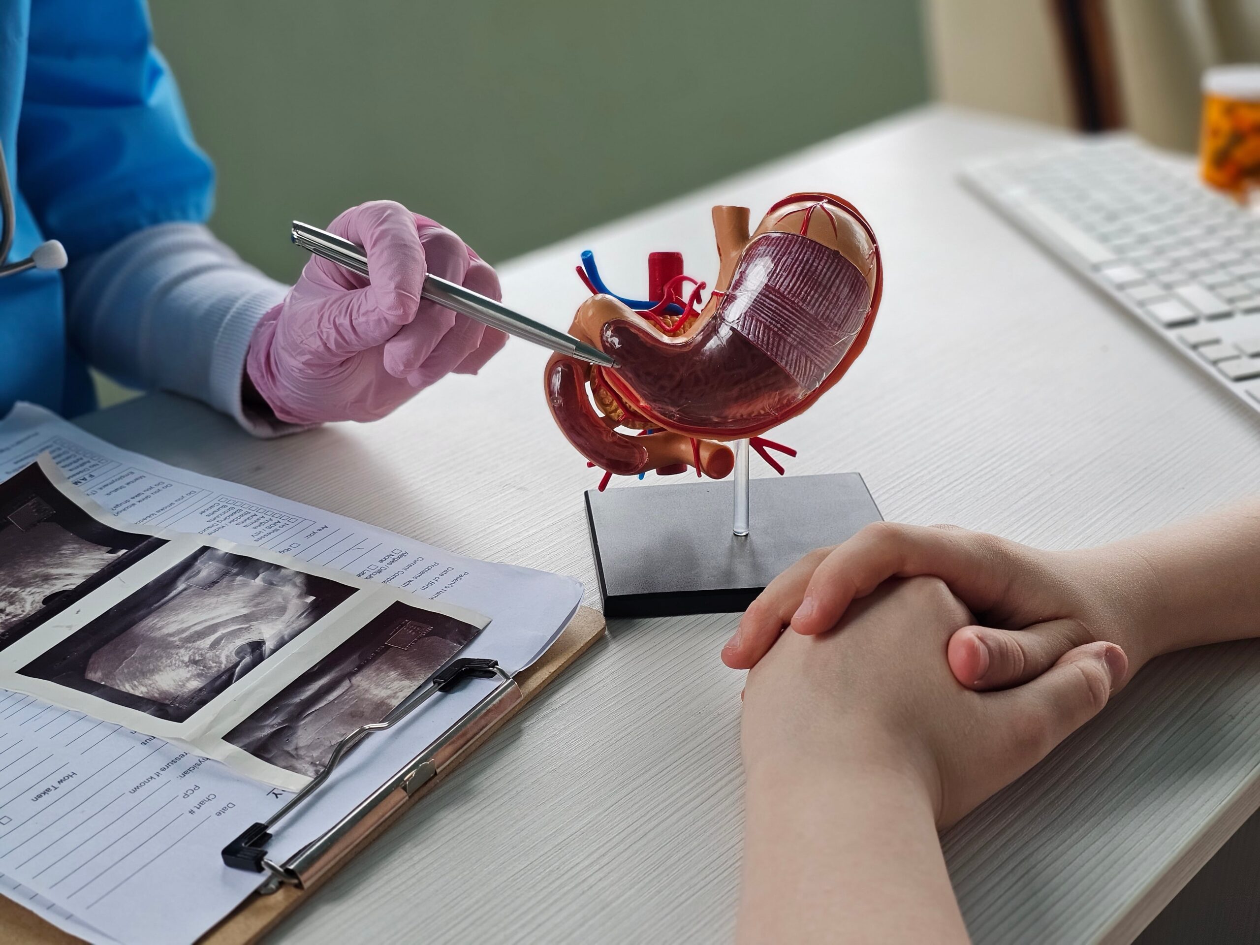 A doctor in blue scrubs points at a stomach model with a pen, talking to a patient. Documents and images rest on a clipboard nearby.