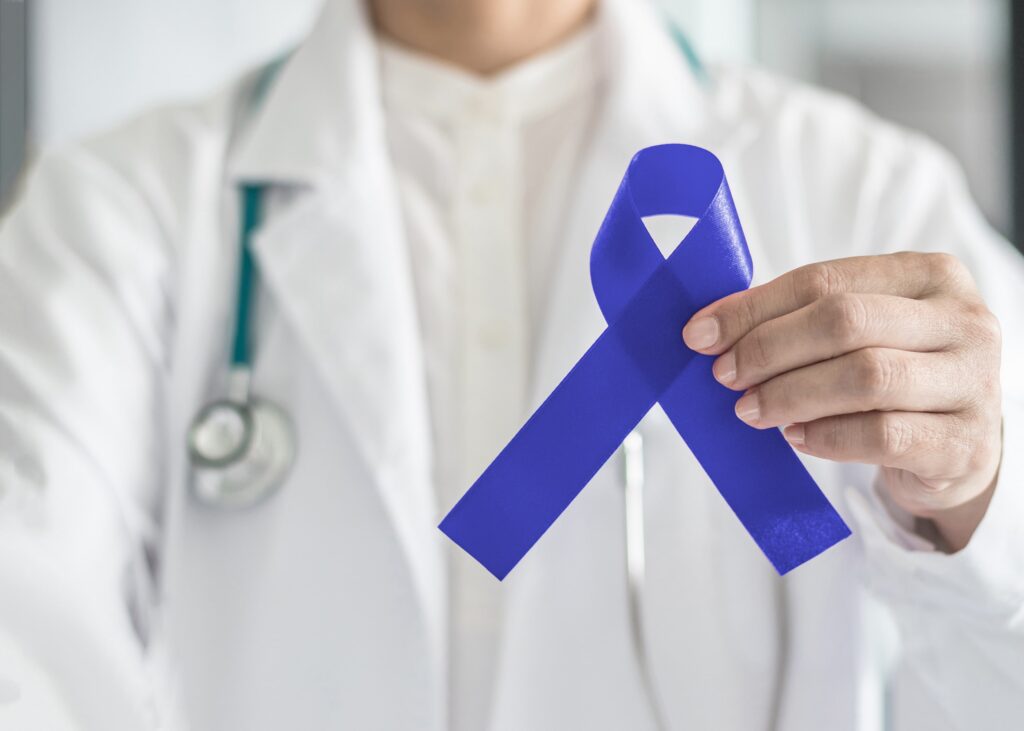 A doctor in a white coat holds a blue awareness ribbon, symbolizing support for health causes. A stethoscope hangs from their neck.