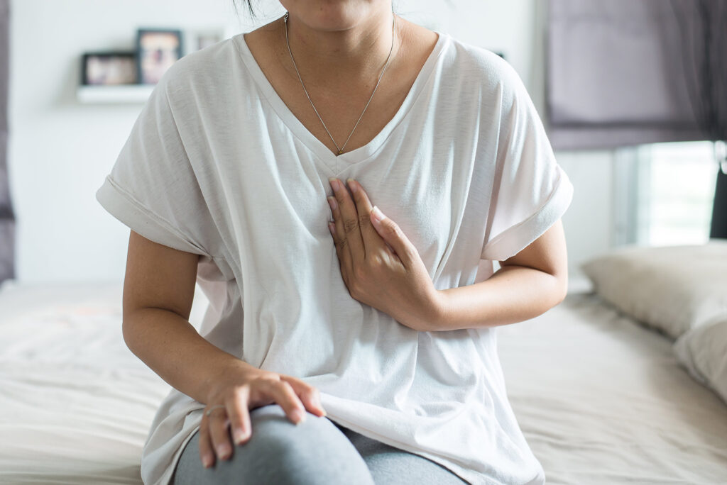 A woman on a bed in a loose white shirt holds her chest with one hand, appearing uncomfortable, possibly from heartburn or pain.
