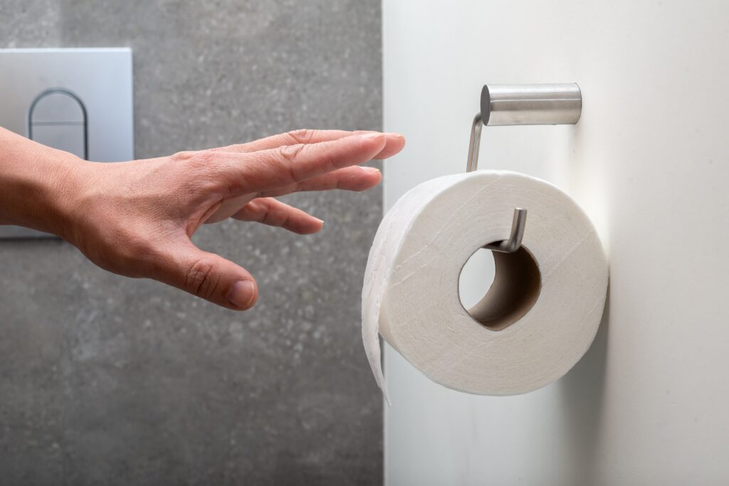 A hand reaches for a roll of toilet paper on a wall in a bathroom with a gray textured wall and flush button in the background.