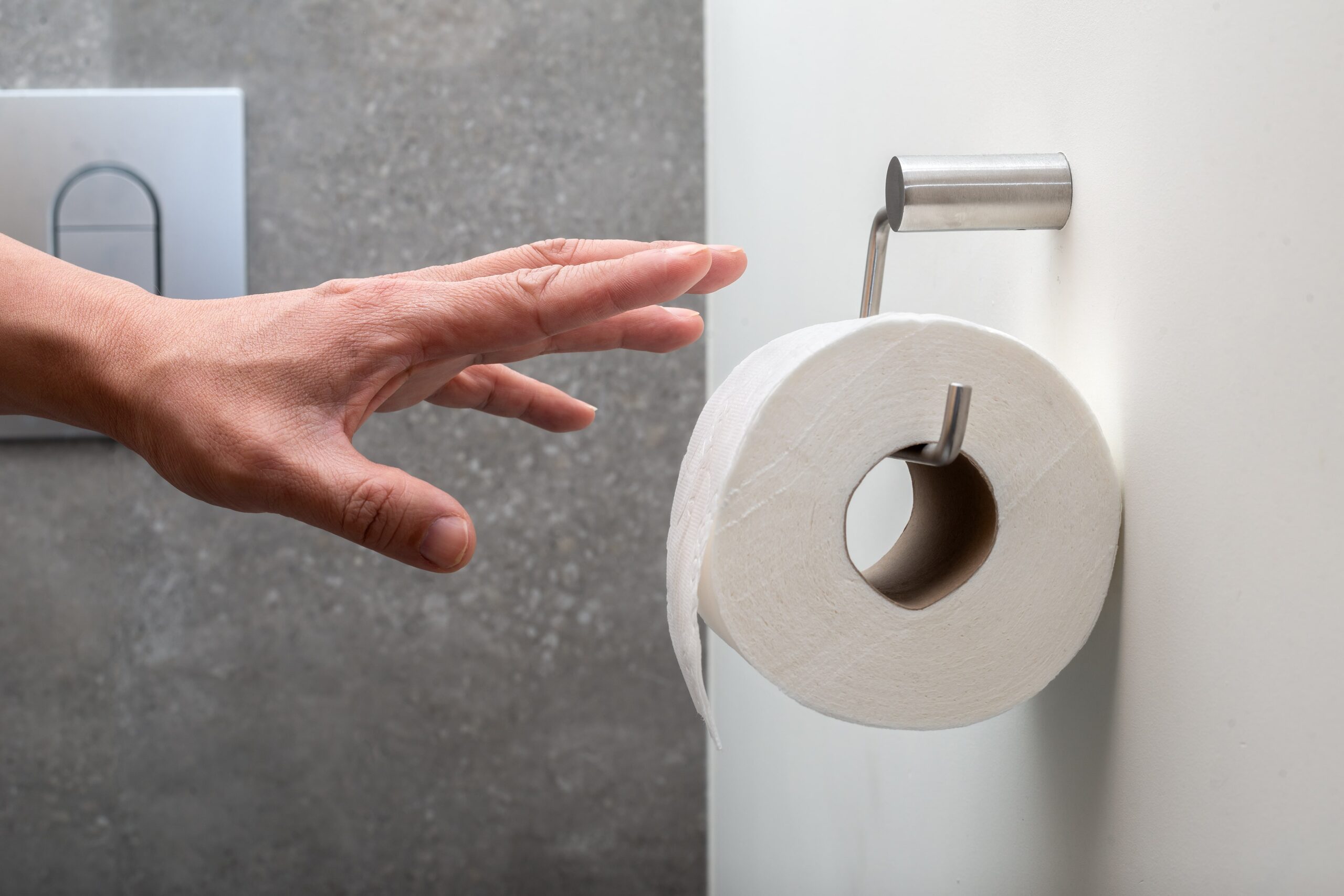 A hand reaches for a roll of toilet paper on a wall in a bathroom with a gray textured wall and flush button in the background.