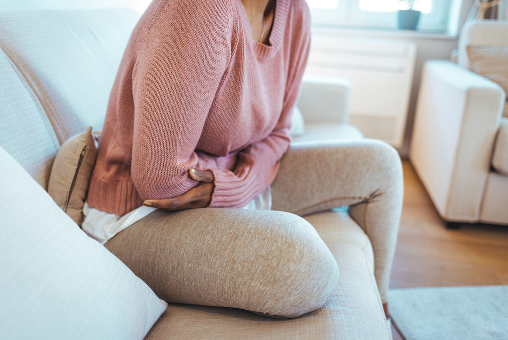 Person on a couch in a pink sweater and beige pants, holding their stomach with a pained look, indicating possible abdominal pain.