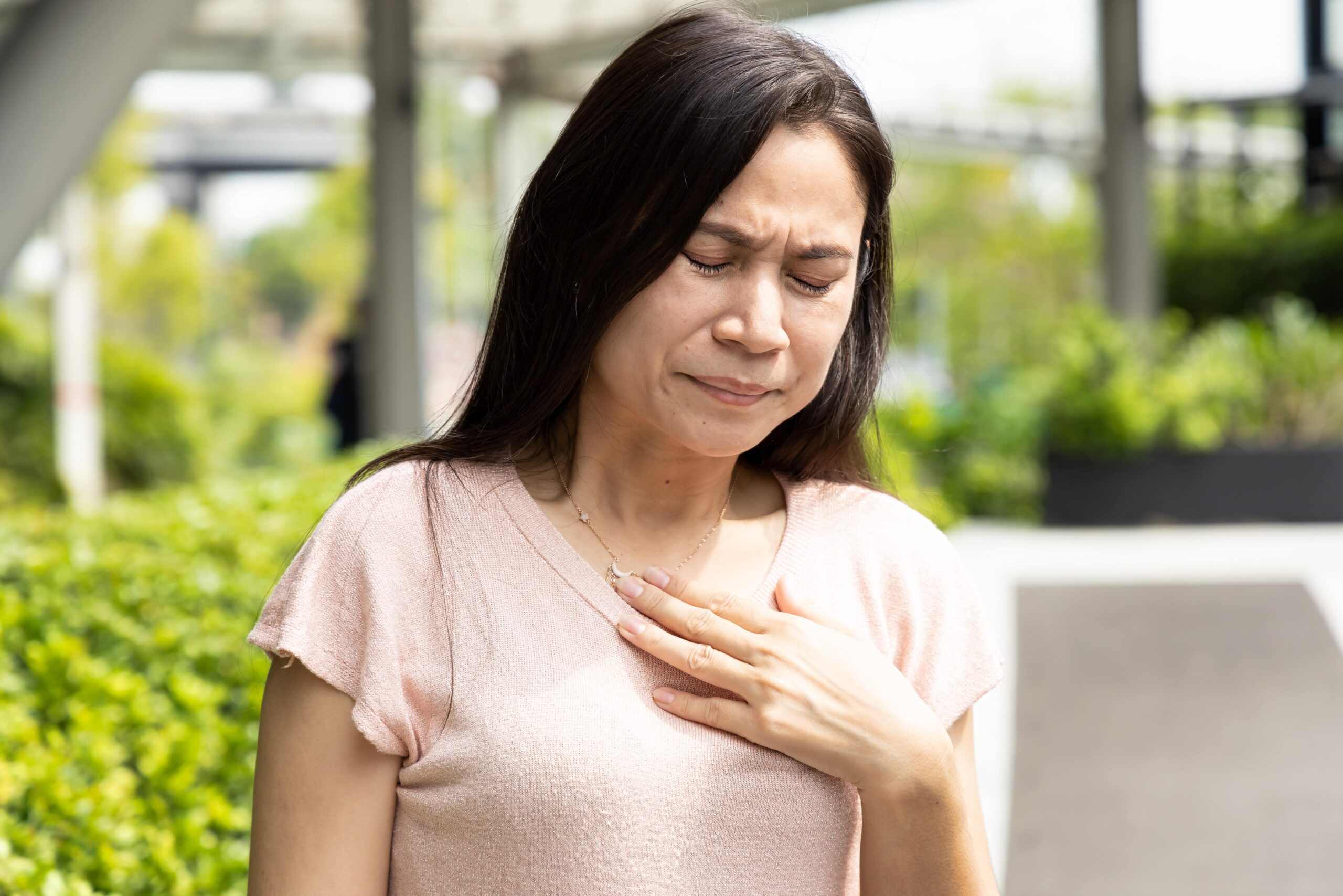 A woman outdoors with eyes closed and one hand on her chest, looking uncomfortable. Greenery and a pathway are in the background.