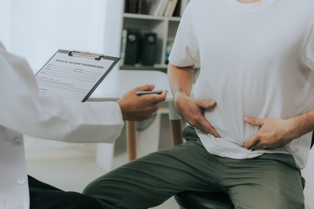 A doctor with a clipboard and pen talks to a patient sitting and touching his lower abdomen, in a medical office setting.