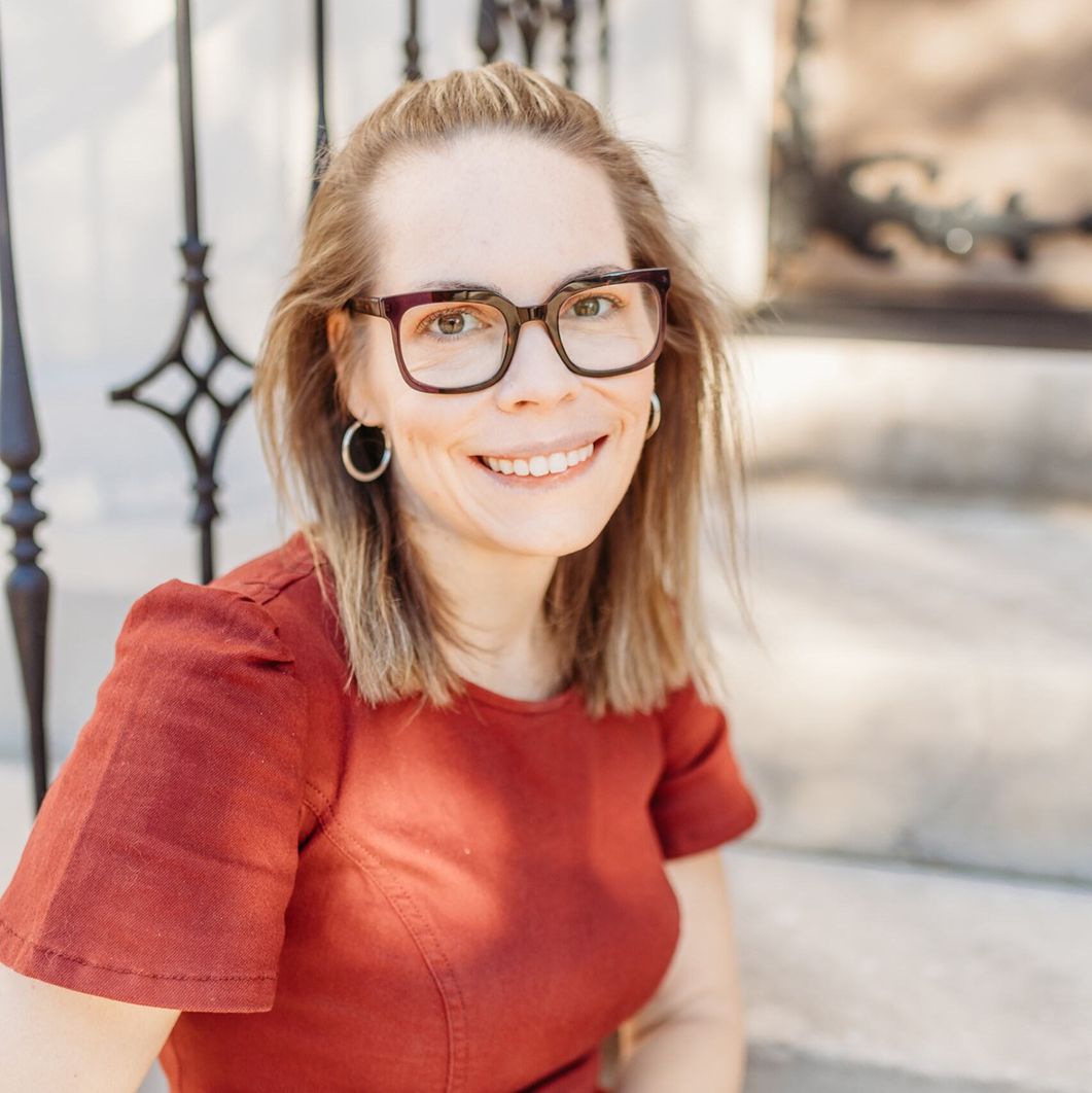 A woman with blonde shoulder-length hair and glasses smiles on outdoor steps, wearing a rust top and hoops; iron railings behind.