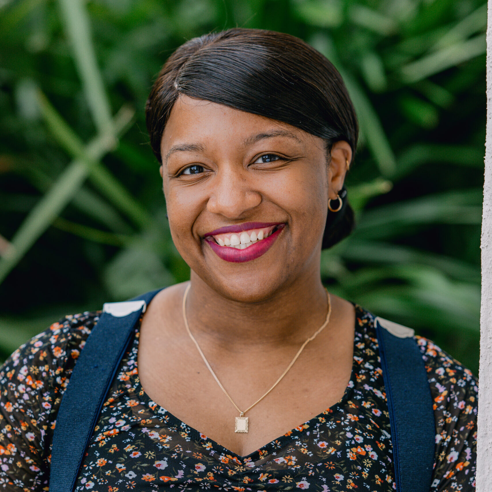 A woman with medium skin, in a floral top, blue suspenders, and gold necklace, smiles with dark hair back before green foliage.