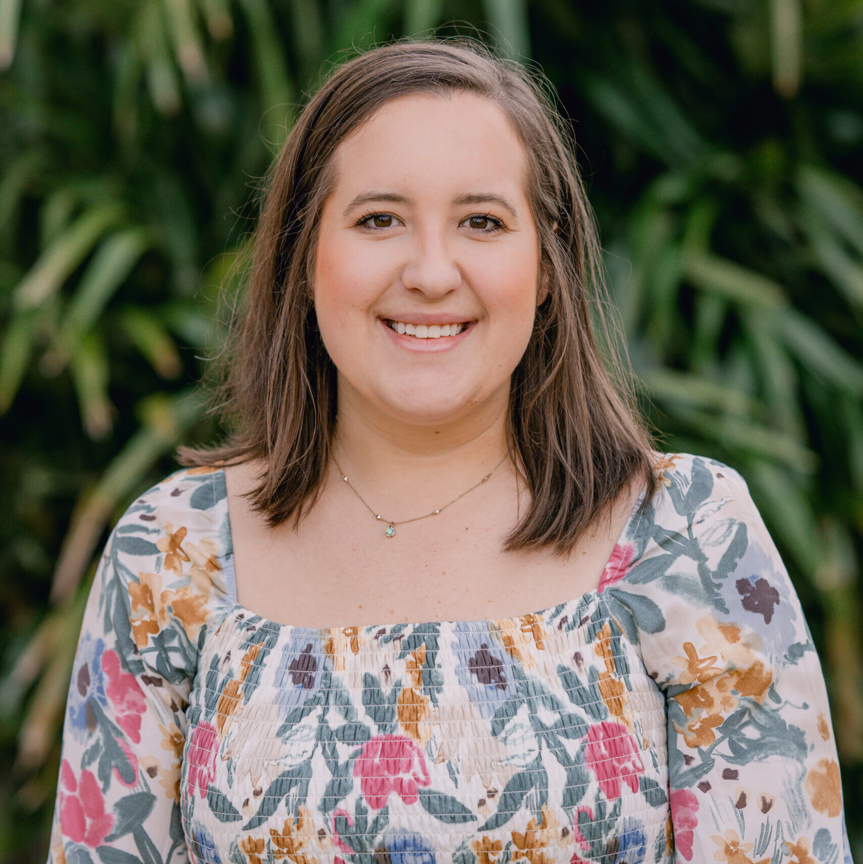 A woman with shoulder-length brown hair smiles at the camera in a floral puff-sleeve dress, standing before lush green foliage.