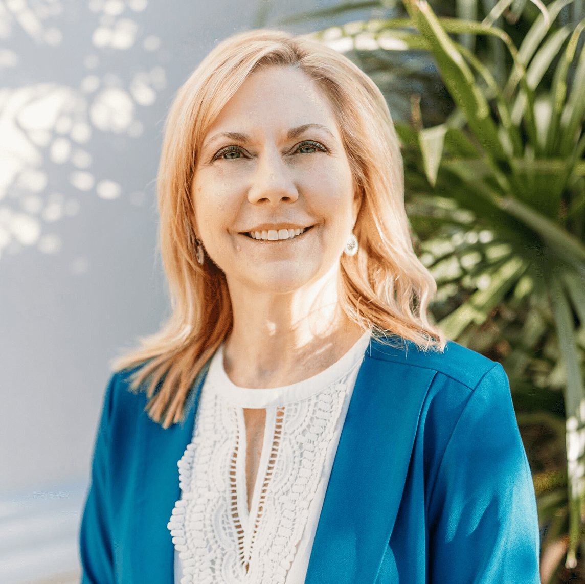 A woman with shoulder-length blonde hair in a blue blazer and white blouse smiles outdoors before green plants and a pale wall.