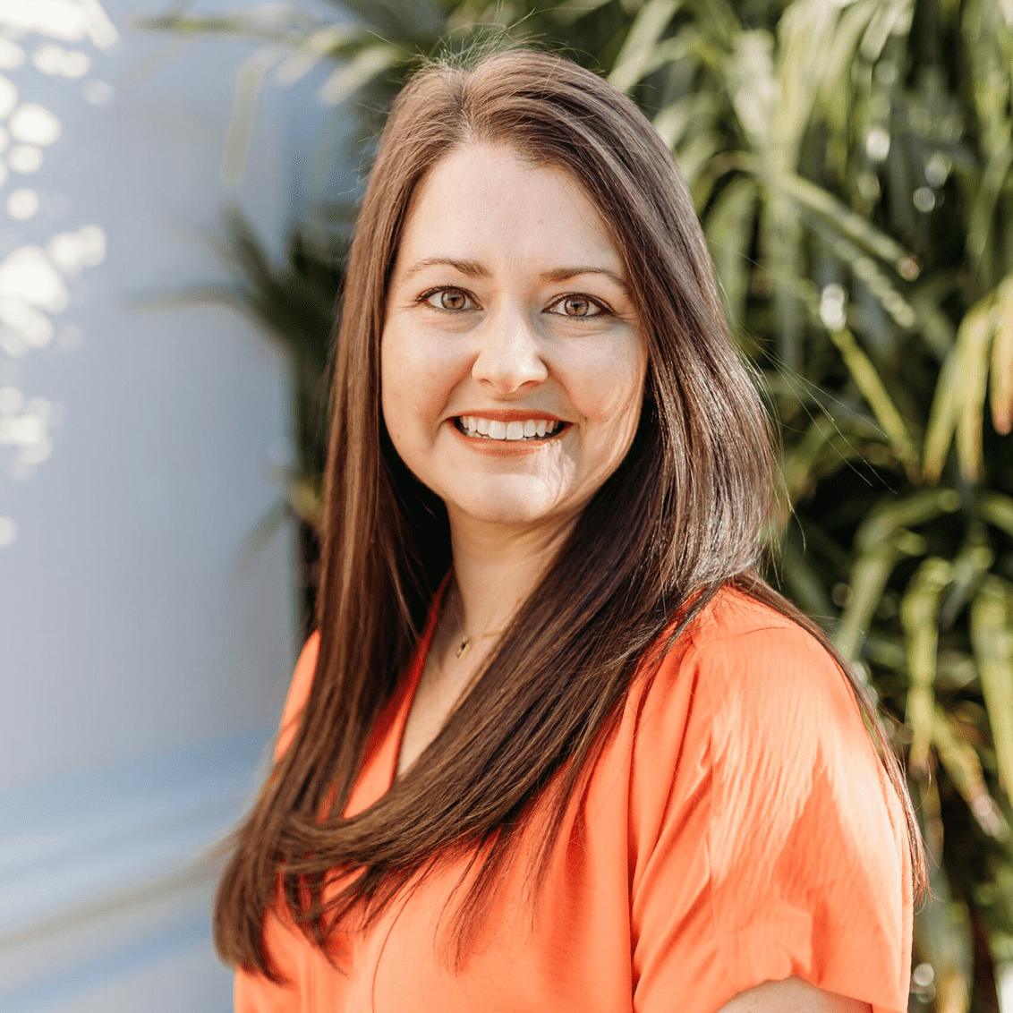 A woman with long brown hair smiles brightly while standing outdoors among green foliage, dressed in an orange blouse and black skirt.
