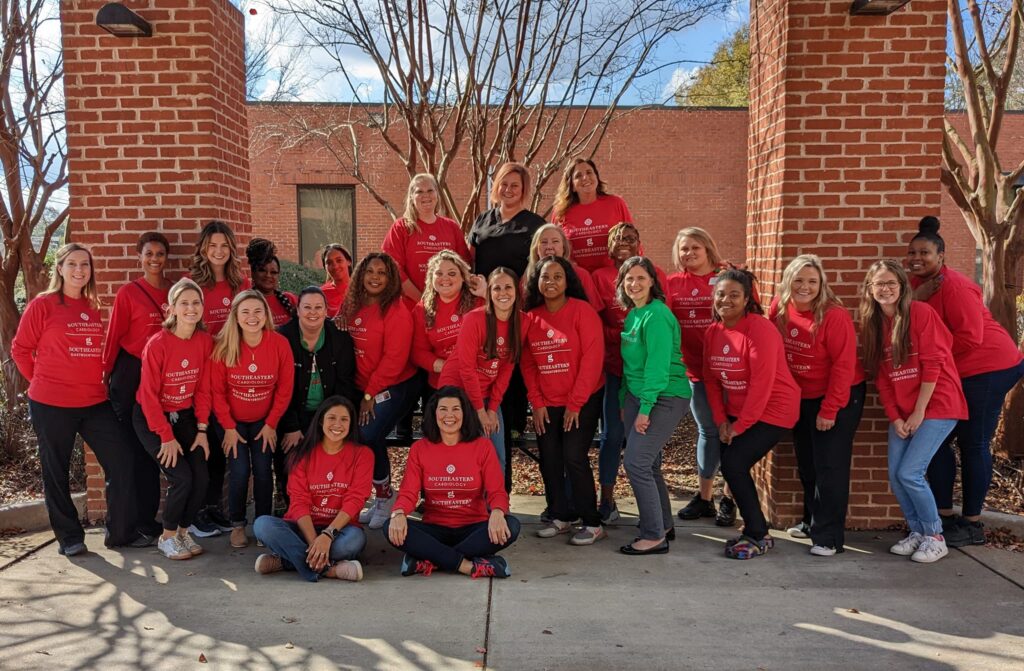 Several women, mainly in matching red shirts with some in green, smile together outdoors by brick columns and bare trees.