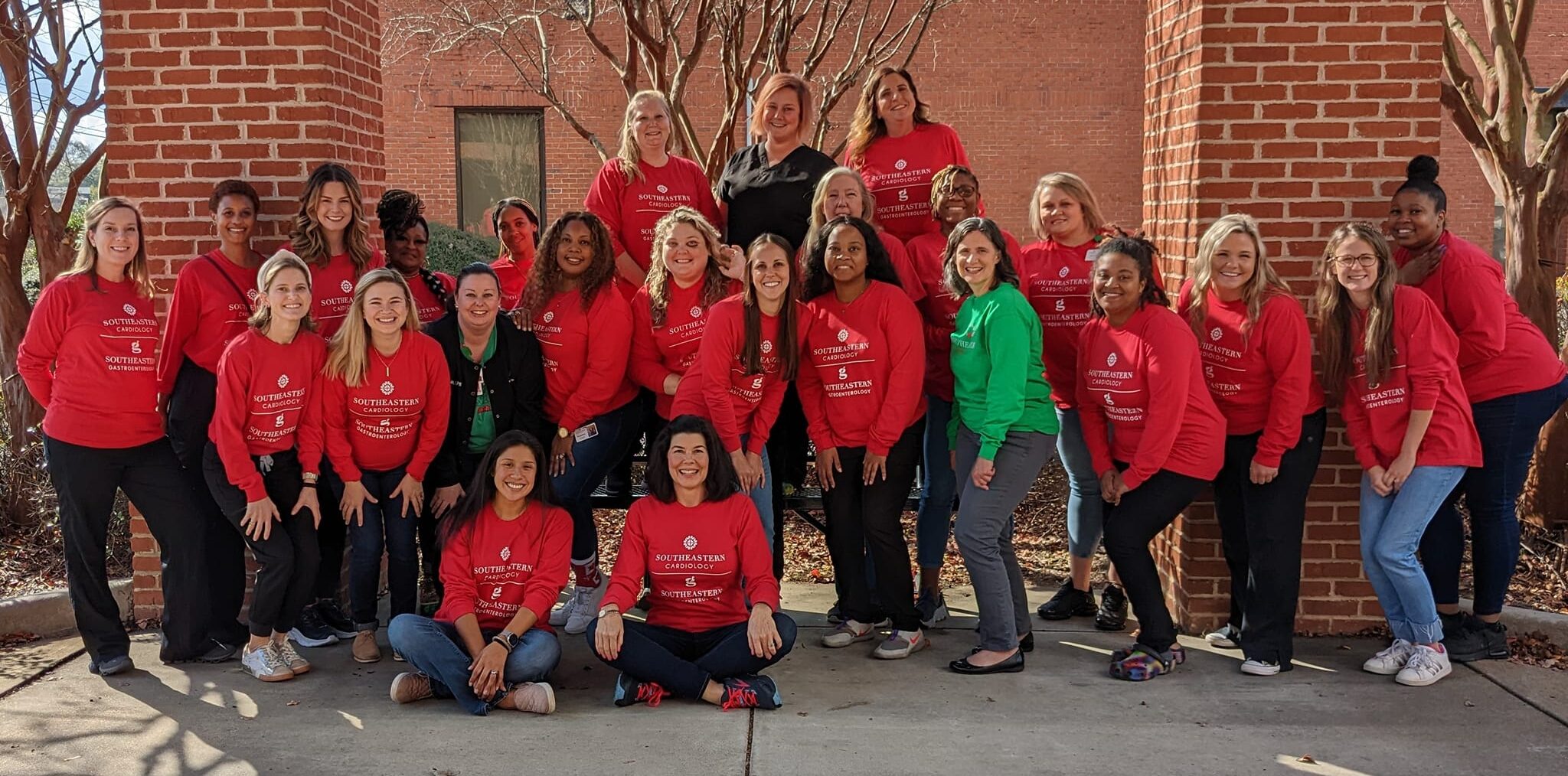 A group of 27 women in matching red shirts poses outdoors by brick columns, with some standing and others sitting; leafless trees behind.
