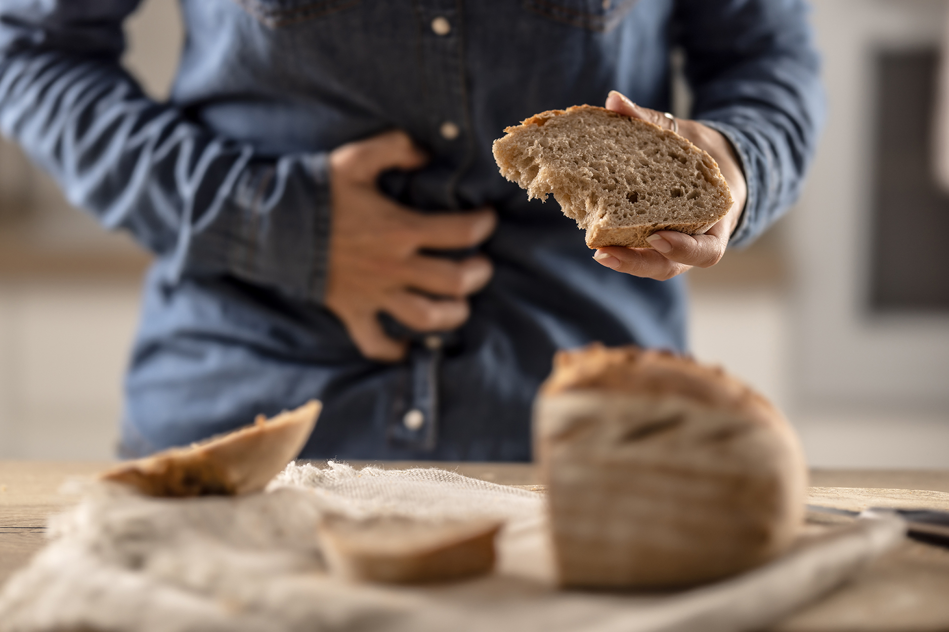 Person holds a slice of bread in one hand, clutching their stomach with the other, looking uncomfortable; bread loaf on cloth nearby.