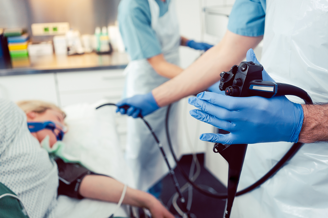 A medical professional in blue gloves holds an endoscope as another assists a patient on a hospital bed in a clinical procedure setting.