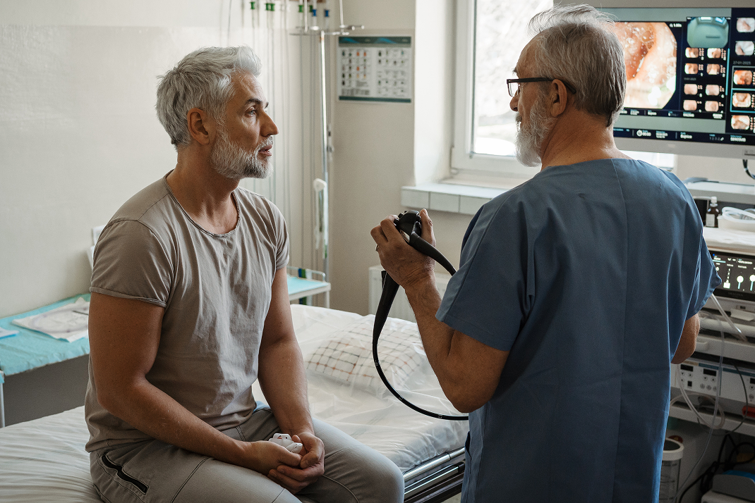 A doctor in blue scrubs holds a medical instrument, talking to a seated male patient in a hospital room with monitors and equipment.