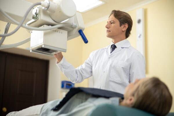 A doctor in a white lab coat uses an X-ray machine above a patient lying on an exam table in a medical clinic.