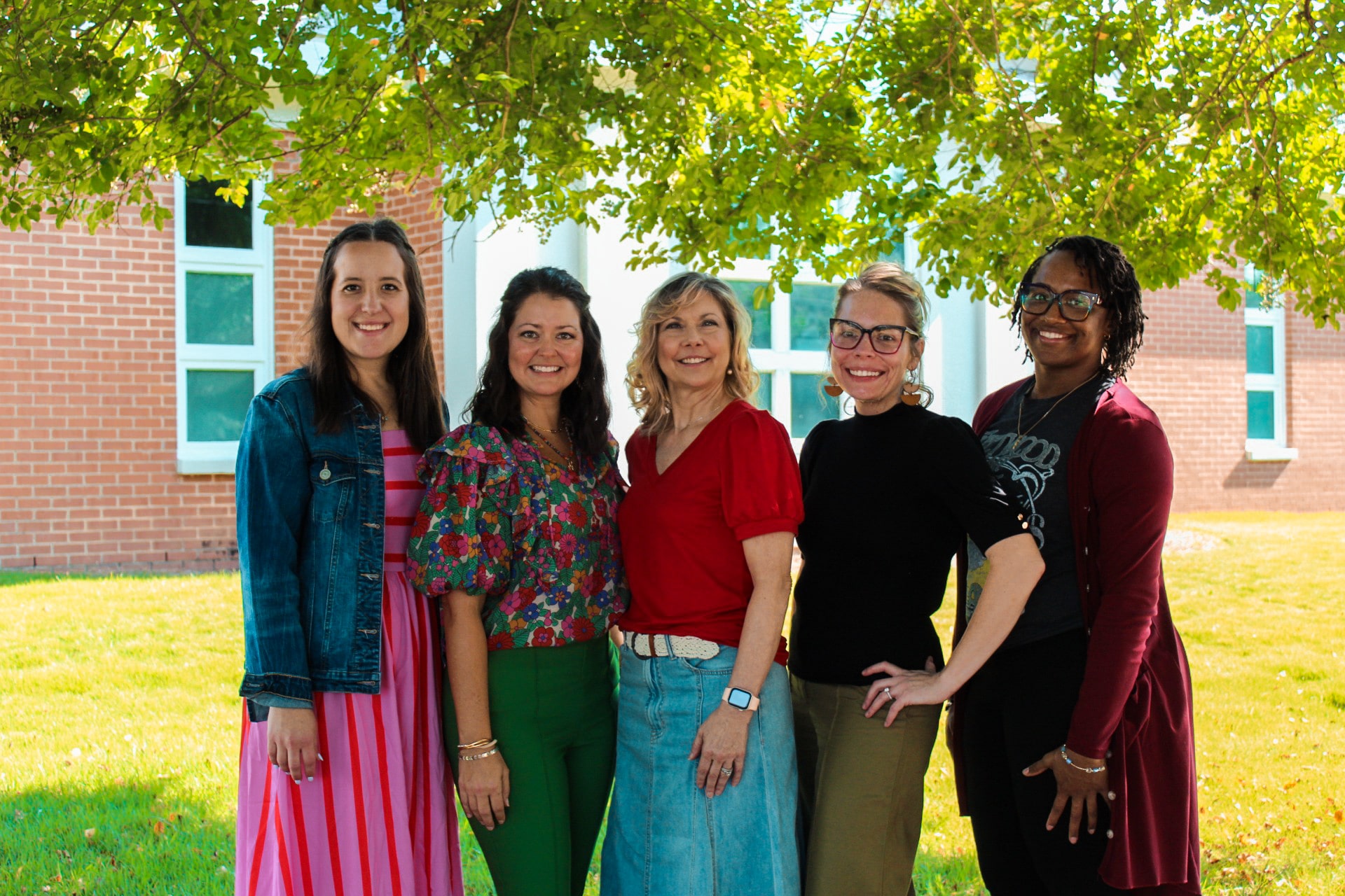 Five women stand outdoors smiling beneath a leafy tree, with dappled sunlight and a brick building in the background.