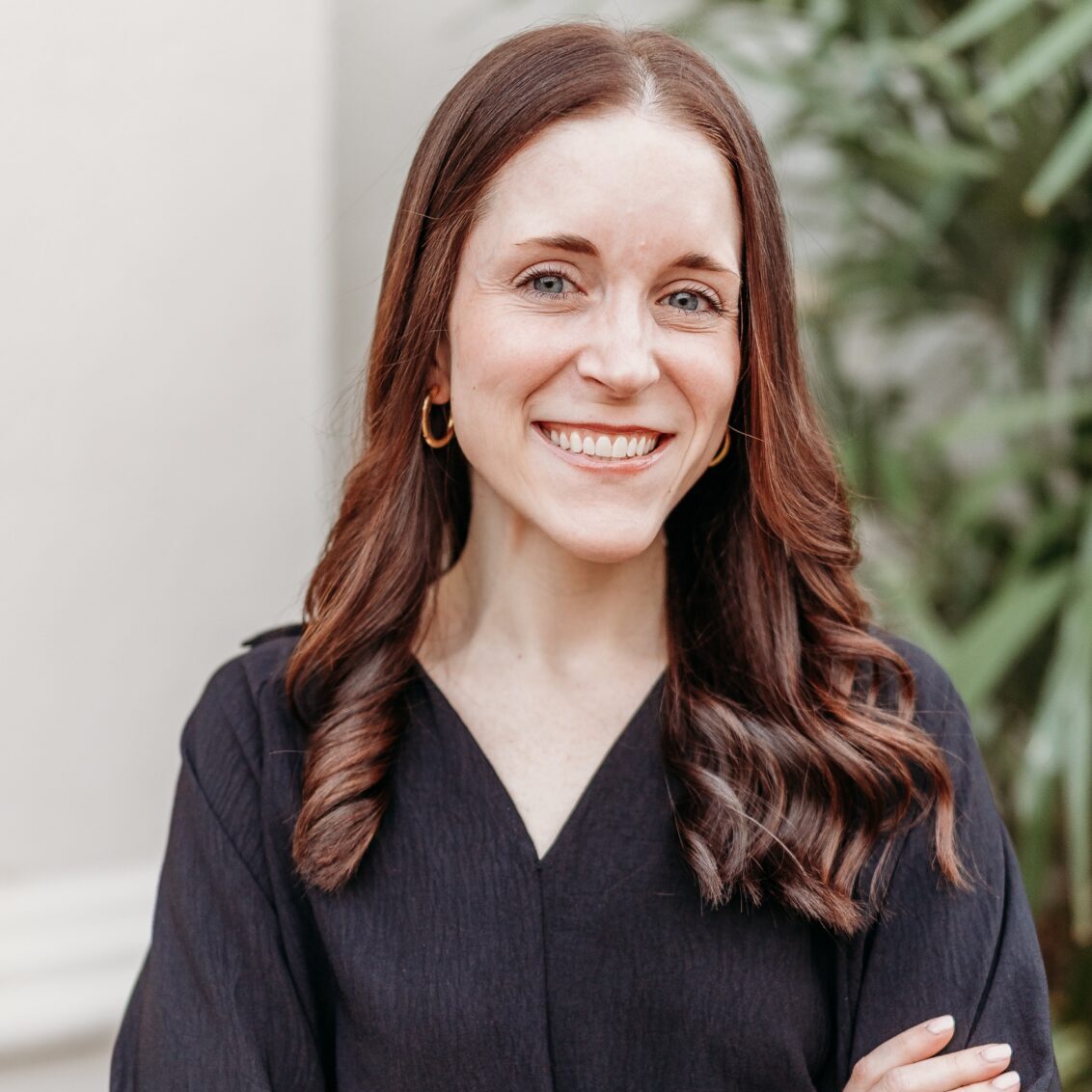 A woman with long brown hair in a black top and gold hoops stands smiling with crossed arms, greenery and a pale wall behind her.