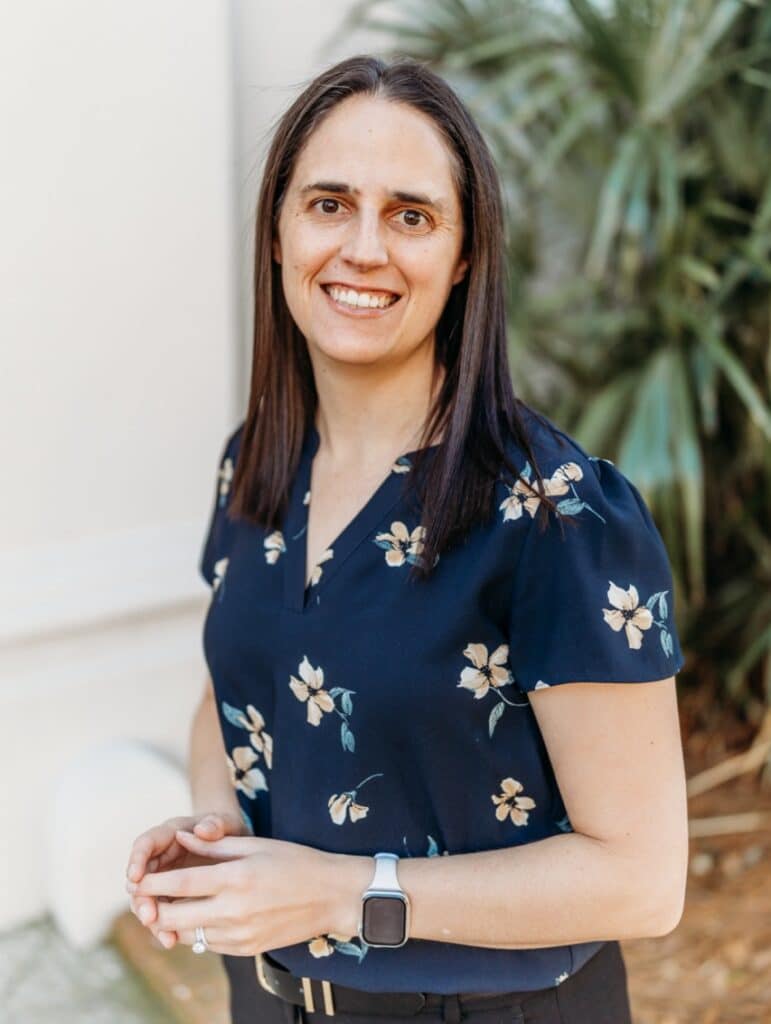 A woman with long brown hair in a navy floral blouse and smartwatch smiles outside by green plants and a light-colored wall.