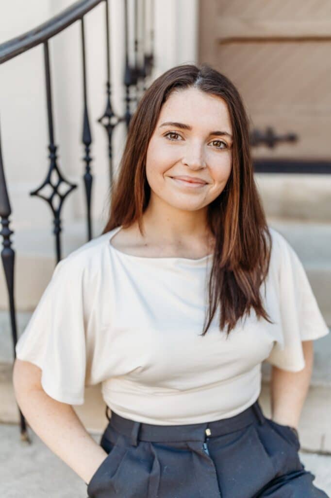 Young woman with long brown hair in a light top smiles on outdoor steps, hand in pocket, iron railings and wooden door behind her.