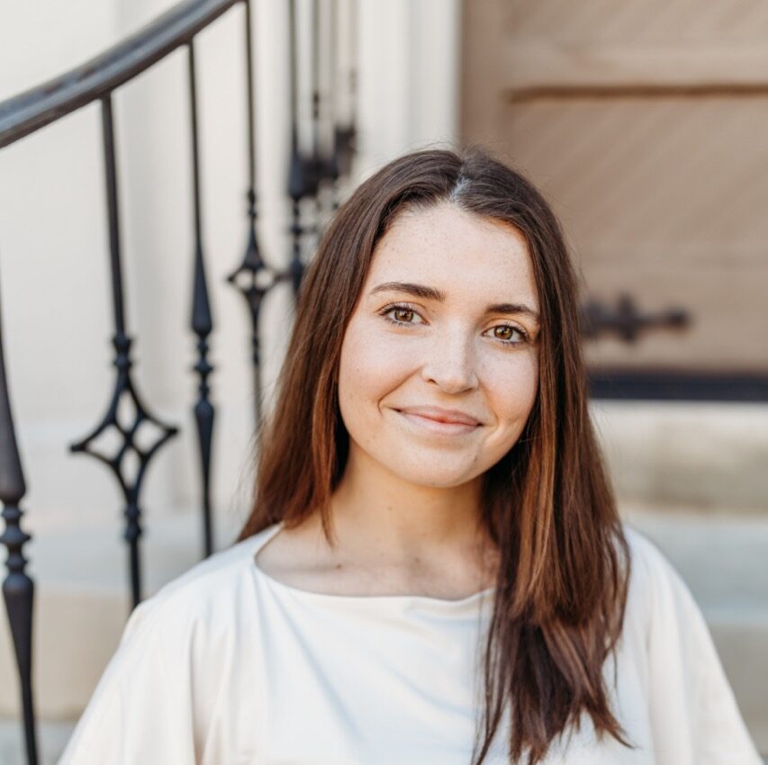 Young woman with long brown hair in a light top smiles on outdoor steps, hand in pocket, iron railings and wooden door behind her.
